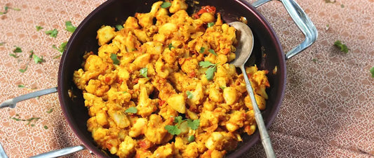 Golden Aloo Gobi curry with cauliflower and potatoes in black pan, garnished with fresh coriander leaves and served with spoon