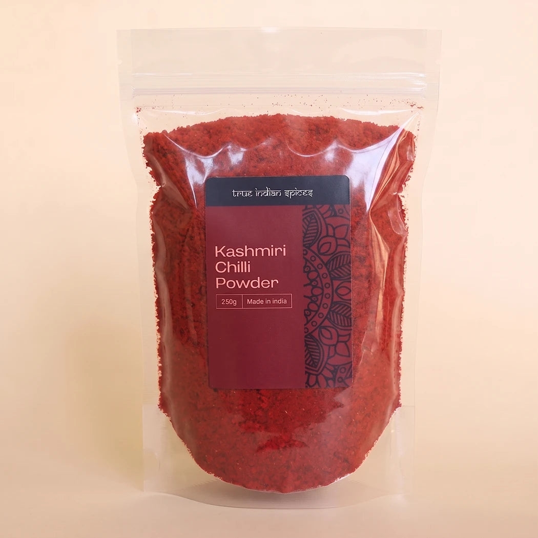 A clear stand-up pouch filled with bright red Kashmiri chili powder, featuring a dark label with white text, set against a solid light beige background.