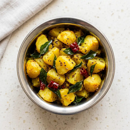 A top-down view of a stainless steel bowl filled with aloo masala in a yellow, garnished with green curry leaves, dried red chilies, and black mustard seeds, set on a light speckled countertop with a folded beige cloth in the corner.