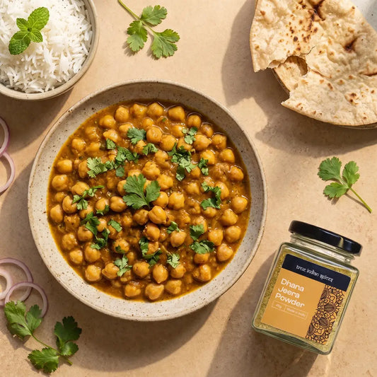 A bowl of chickpea curry with green herbs on top, served with white rice, flatbread, and onion slices on a beige surface, with a jar of True Indian Spicse powder in the foreground.
