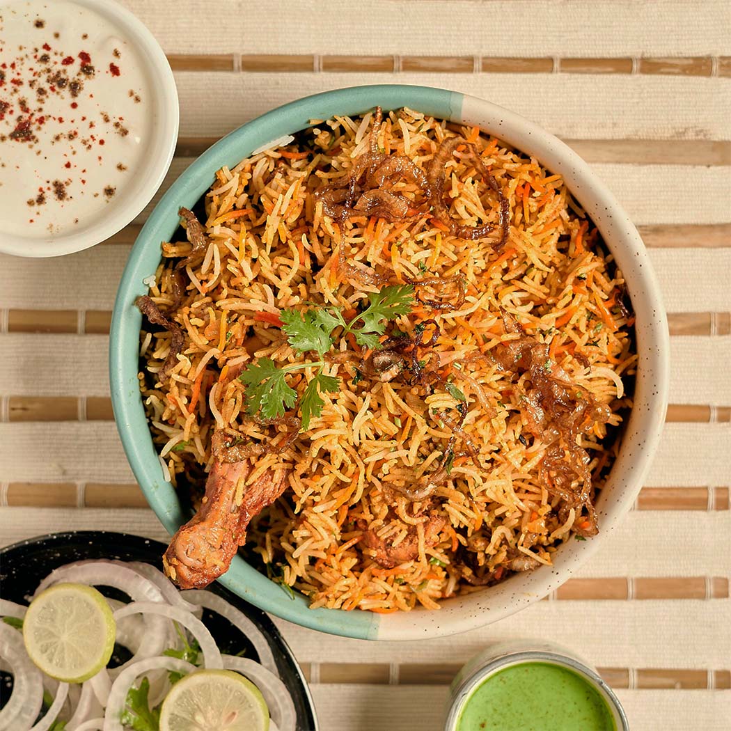 A bowl chicken biryani topped with fresh coriander leaves and fried onions, accompanied by a side of spiced yogurt, sliced onions, lemon wedges, and a glass of green chutney.