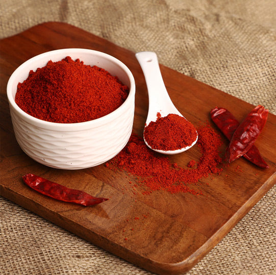 White ceramic bowl filled with red Kashmiri chilli powder on wooden cutting board, with measuring spoon and dried red chilies as props on burlap background