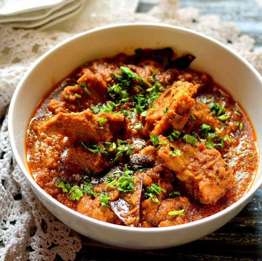 Traditional Indian curry dish rogan josh in white bowl featuring tender meat pieces in rich red sauce, garnished with fresh green herbs