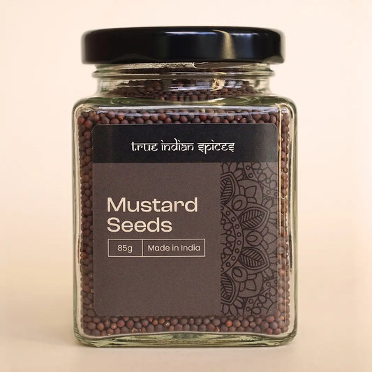 A clear glass jar of mustard seeds with a black lid and a dark brown label reading 'true indian spices' and 'Mustard Seeds 85g Made in India', set against a plain light beige background.
