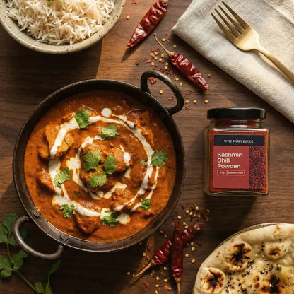 Butter chicken with naan bread, rice, and Kashmiri Chilli Powder jar on a wooden table.