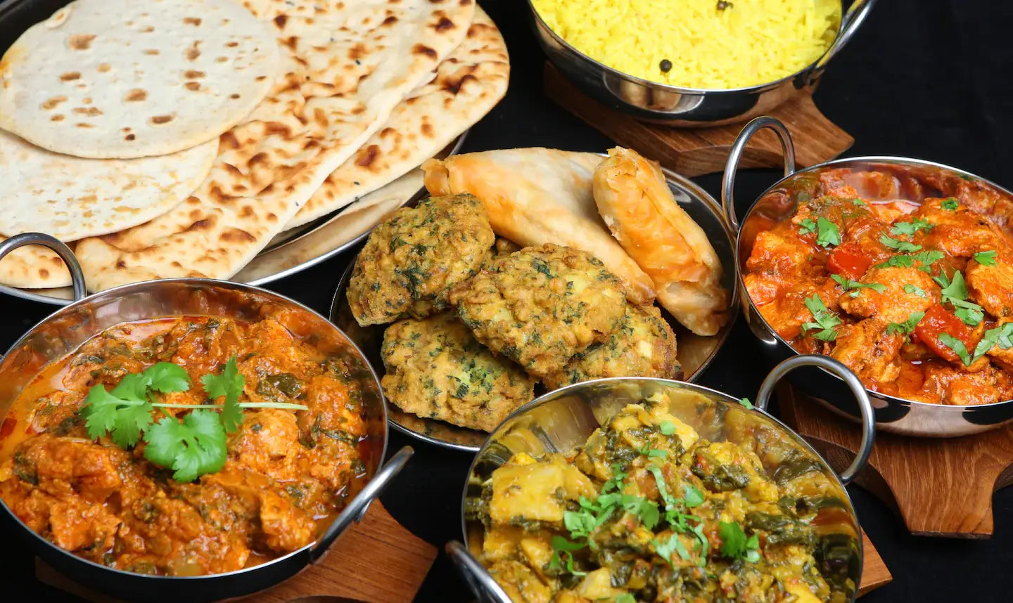 Delicious Indian dishes arranged on a table, featuring naan, yellow rice, and a variety of curries, samosas, and pakoras. Showcasing the vibrant, rich flavours of Indian cuisine.