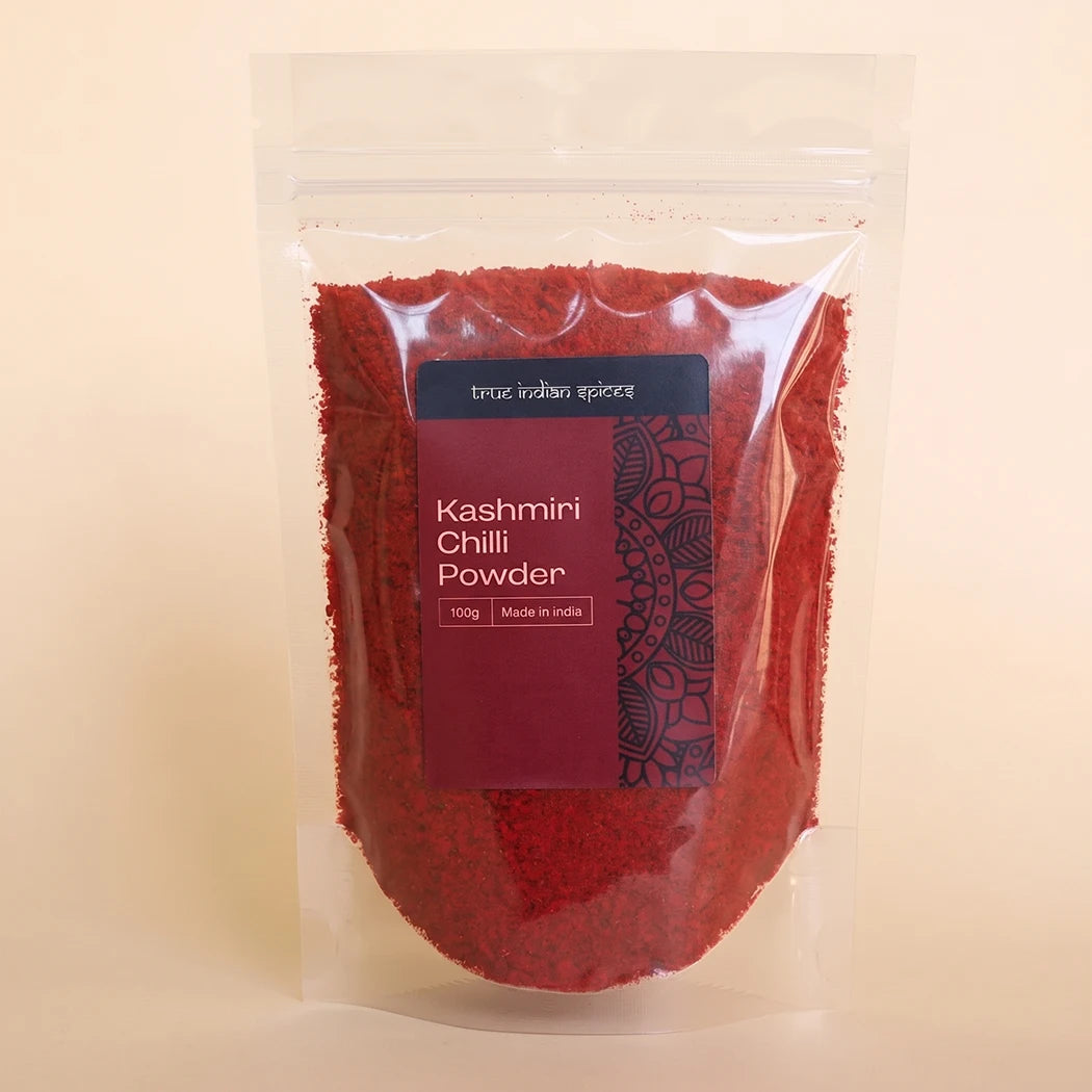 A clear stand-up pouch filled with bright red Kashmiri chili powder, featuring a dark red label with white text and a black mandala design, set against a solid light beige background.