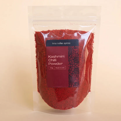 A clear stand-up pouch filled with bright red Kashmiri chili powder, featuring a dark red label with white text and a black mandala design, set against a solid light beige background.