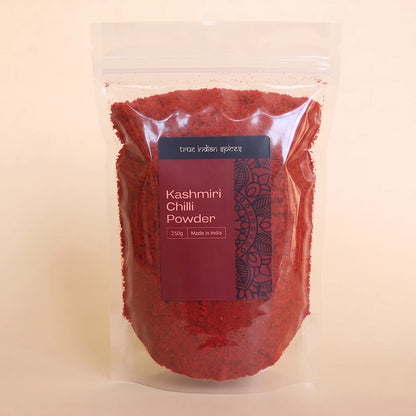 A clear stand-up pouch filled with bright red Kashmiri chili powder, featuring a dark label with white text, set against a solid light beige background.