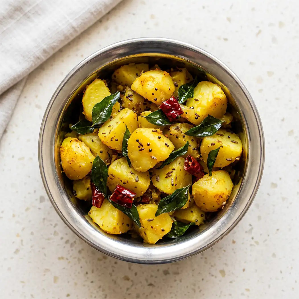 A top-down view of a stainless steel bowl filled with aloo masala in a yellow, garnished with green curry leaves, dried red chilies, and black mustard seeds, set on a light speckled countertop with a folded beige cloth in the corner.