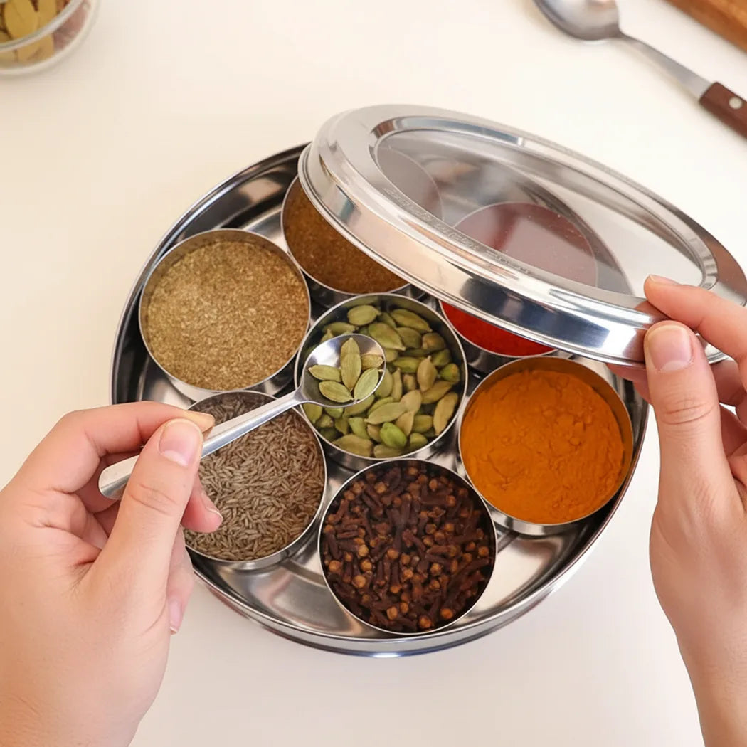 Indian spice box with various spices and a hand holding a spoon.